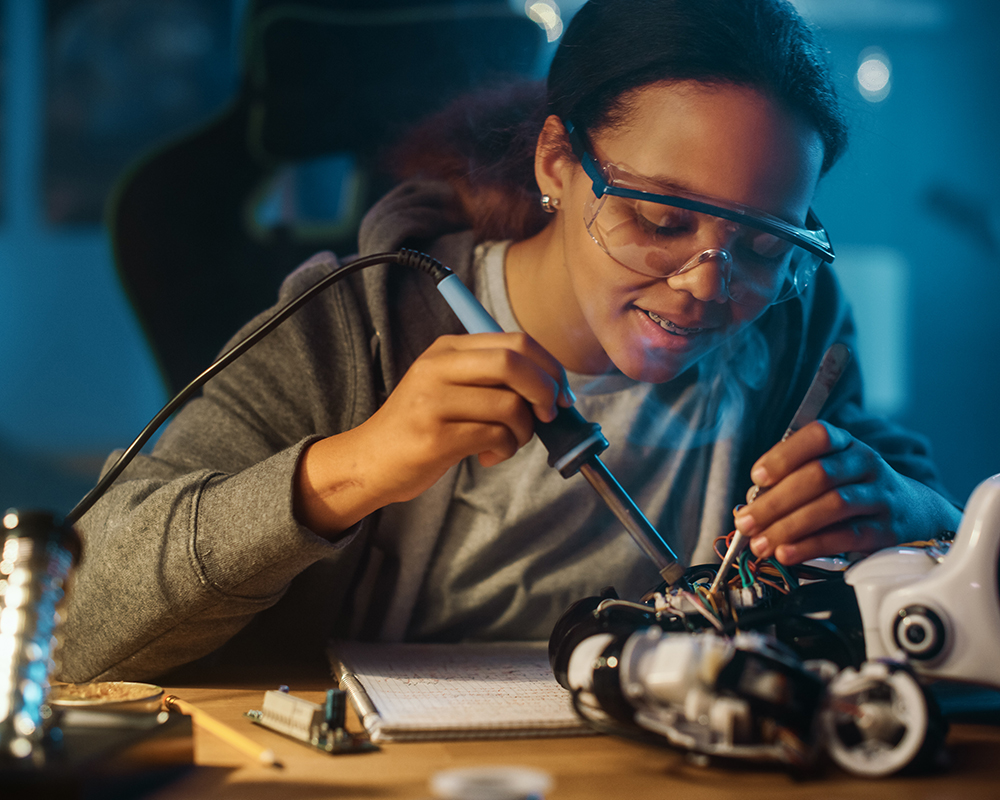 Young Multiethnic Woman is Studying Electronics and Soldering Wires and Circuit Boards in Her Science Robotics Project. Woman is Working on a Robot.